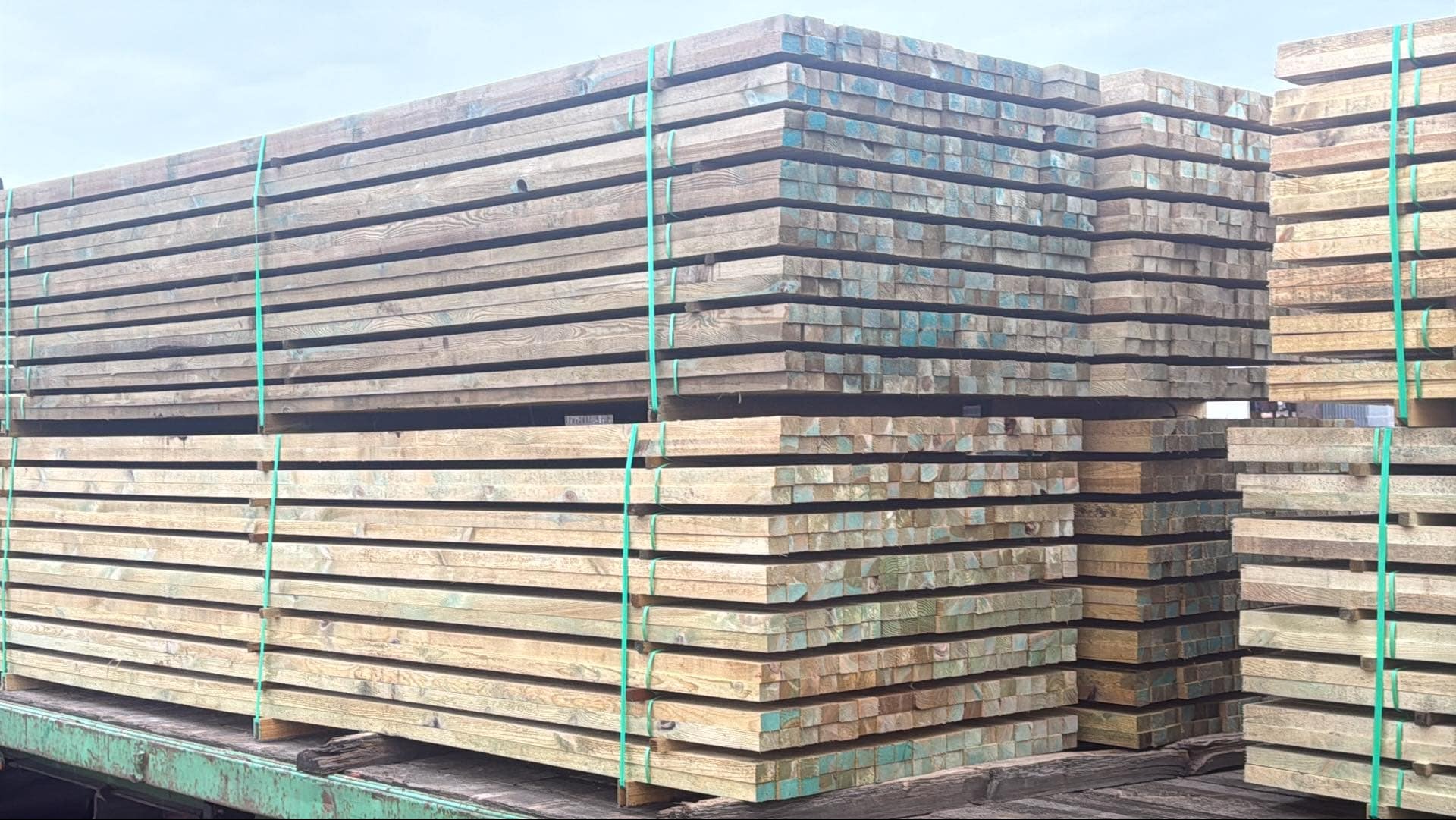 Stosy impregnowanych desek na paletach, zabezpieczone zielonymi taśmami. Widoczne ślady impregnacji na końcach desek.