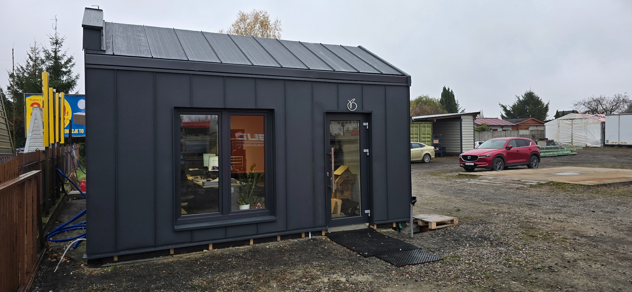 Nowoczesny, grafitowy budynek biurowy z blachy trapezowej, z oknem i drzwiami, na tle placu z zaparkowanymi samochodami. Widoczny fragment ogrodzenia i reklamy.