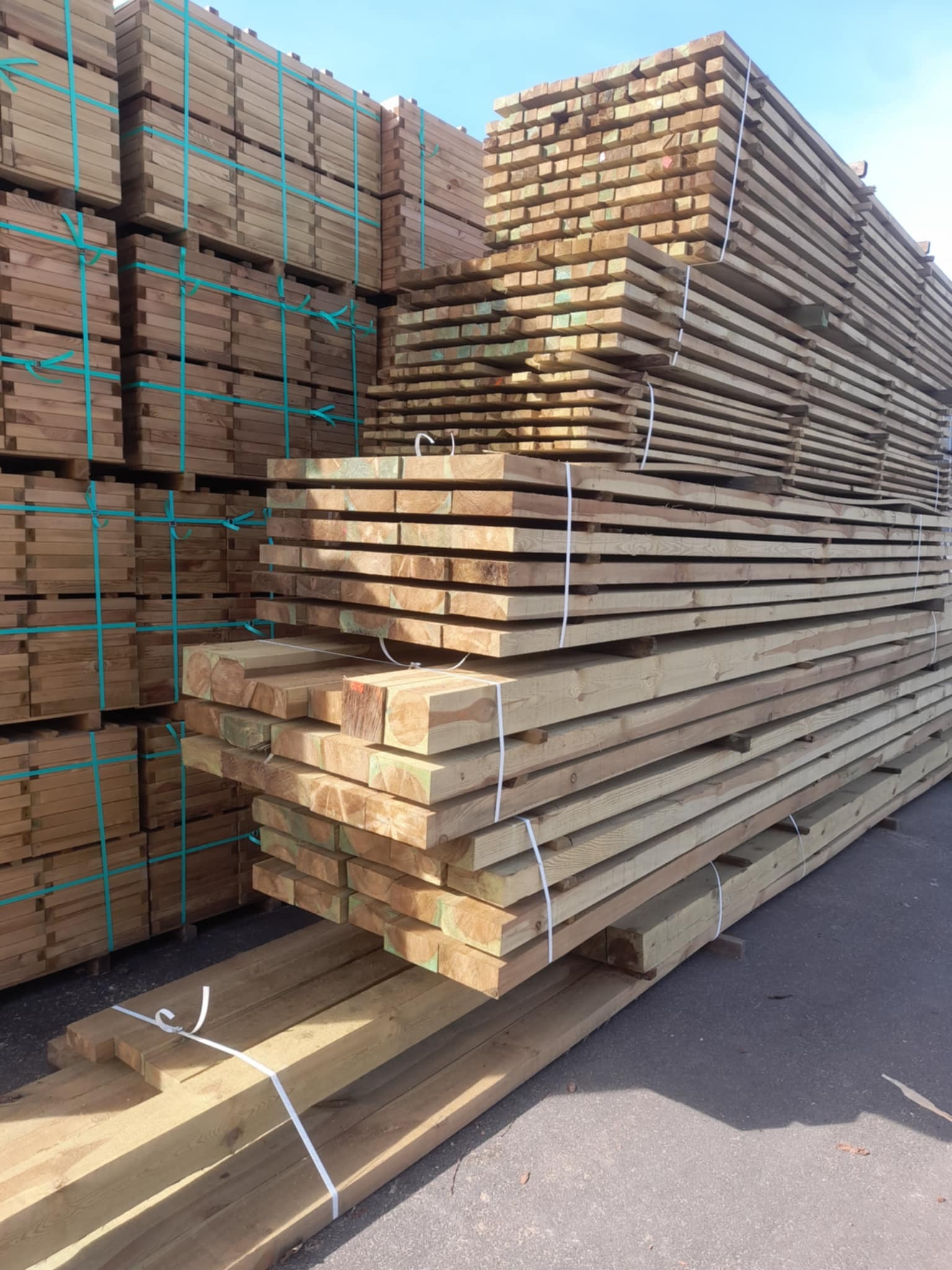 Stosy tarcicy budowlanej, zabezpieczone taśmami, ułożone na placu składowym pod błękitnym niebem. Widoczne różne przekroje i długości drewna.