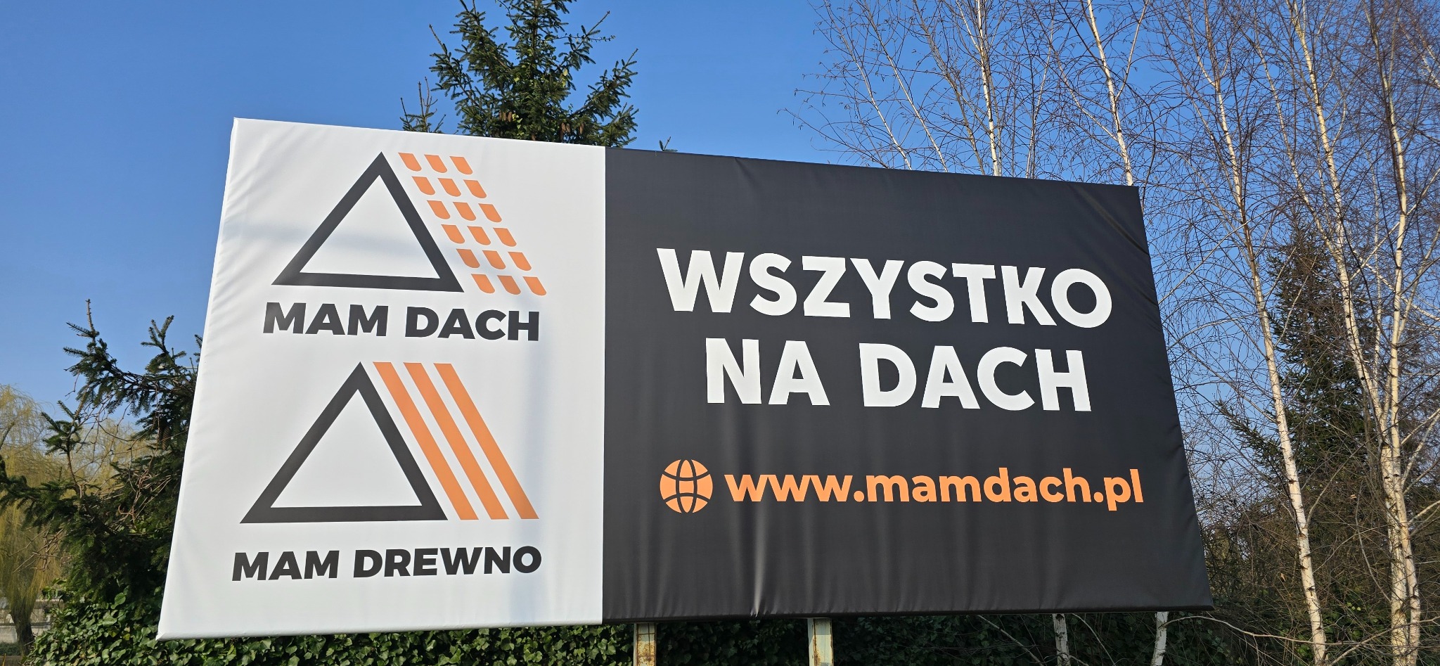 Billboard reklamowy firmy 'MAM DACH' z hasłem 'Wszystko na dach' na tle błękitnego nieba i drzew. Widoczne logo firmy i adres strony internetowej.
