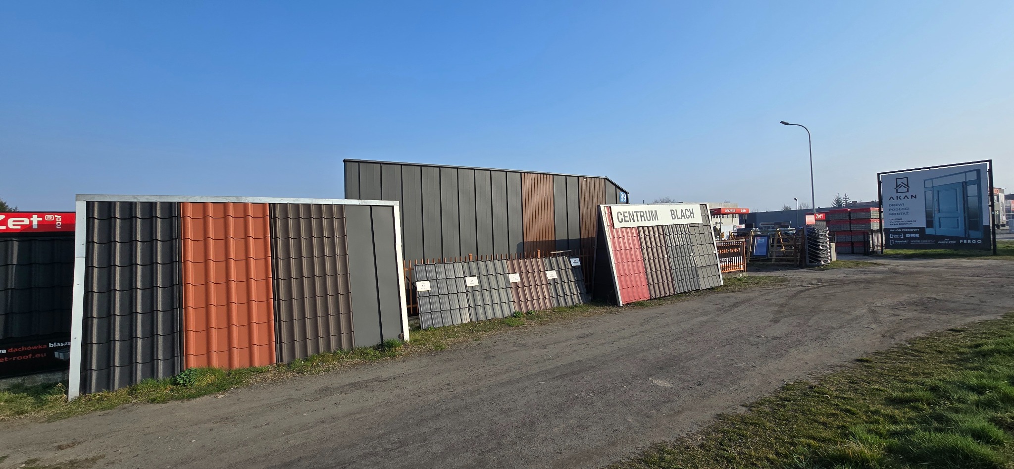 Ekspozycja różnokolorowych dachówek i blach trapezowych na zewnątrz, prezentacja materiałów budowlanych w składzie z widocznym szyldem 'Centrum Blach'.