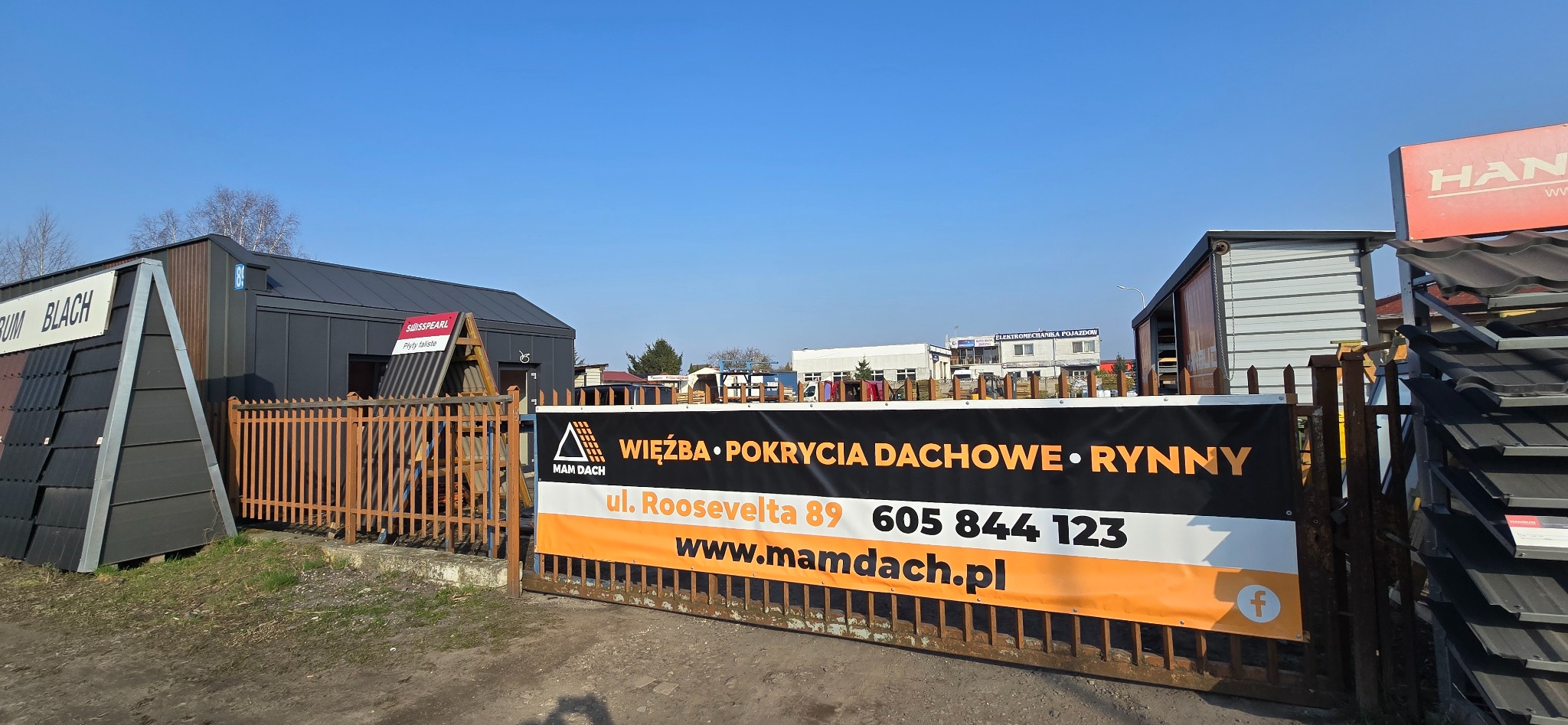 Ekspozycja materiałów dachowych: blacha, dachówki i rynny. Baner reklamowy firmy 'Mam Dach' na ogrodzeniu. Widoczne budynki i elementy ekspozycji.
