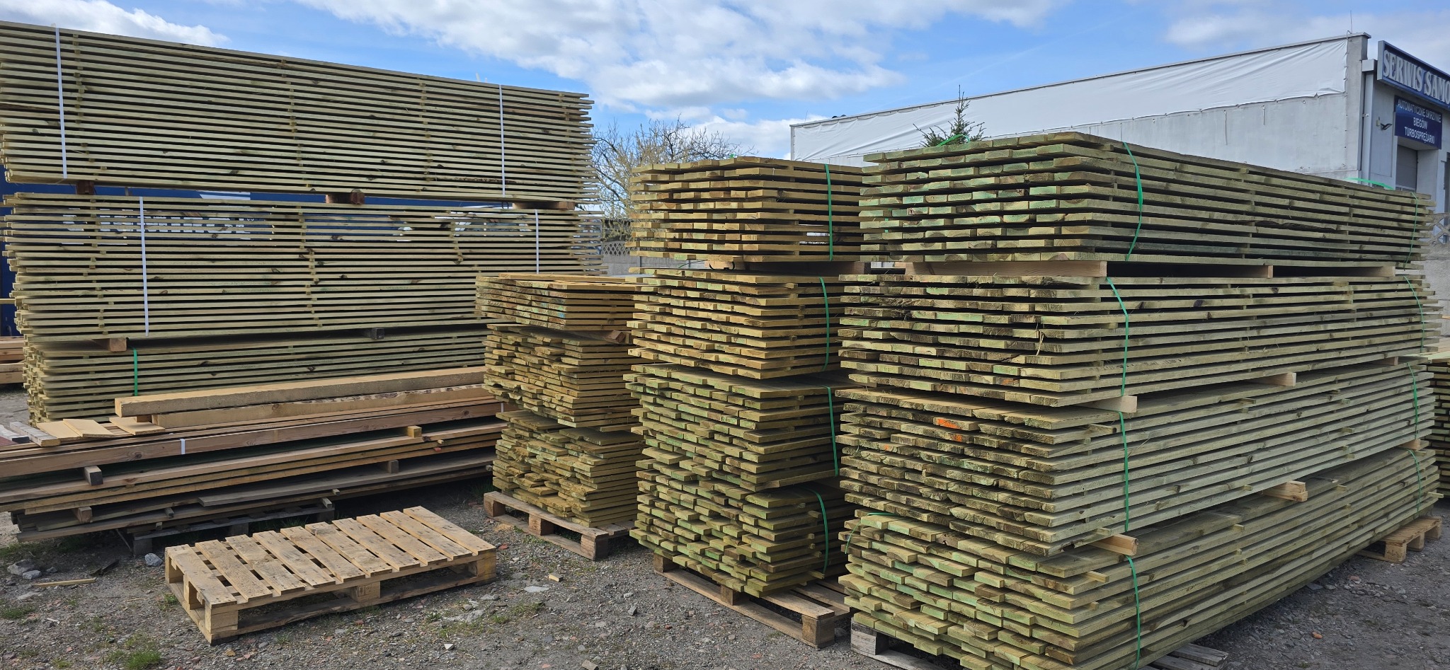 Stosy impregnowanego drewna budowlanego, ułożone na paletach, widoczne na placu składowym pod błękitnym niebem z chmurami. Drewno zabezpieczone taśmami.