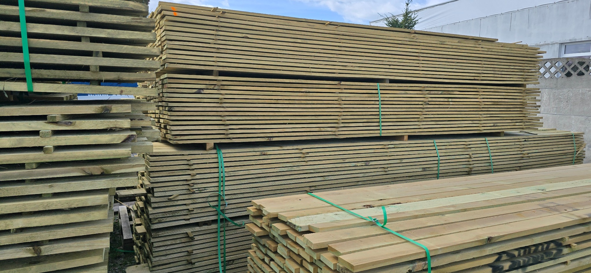 Stosy impregnowanego drewna budowlanego, zabezpieczone zieloną taśmą, na tle betonowego muru i fragmentu budynku. Widoczne deski ułożone warstwami.