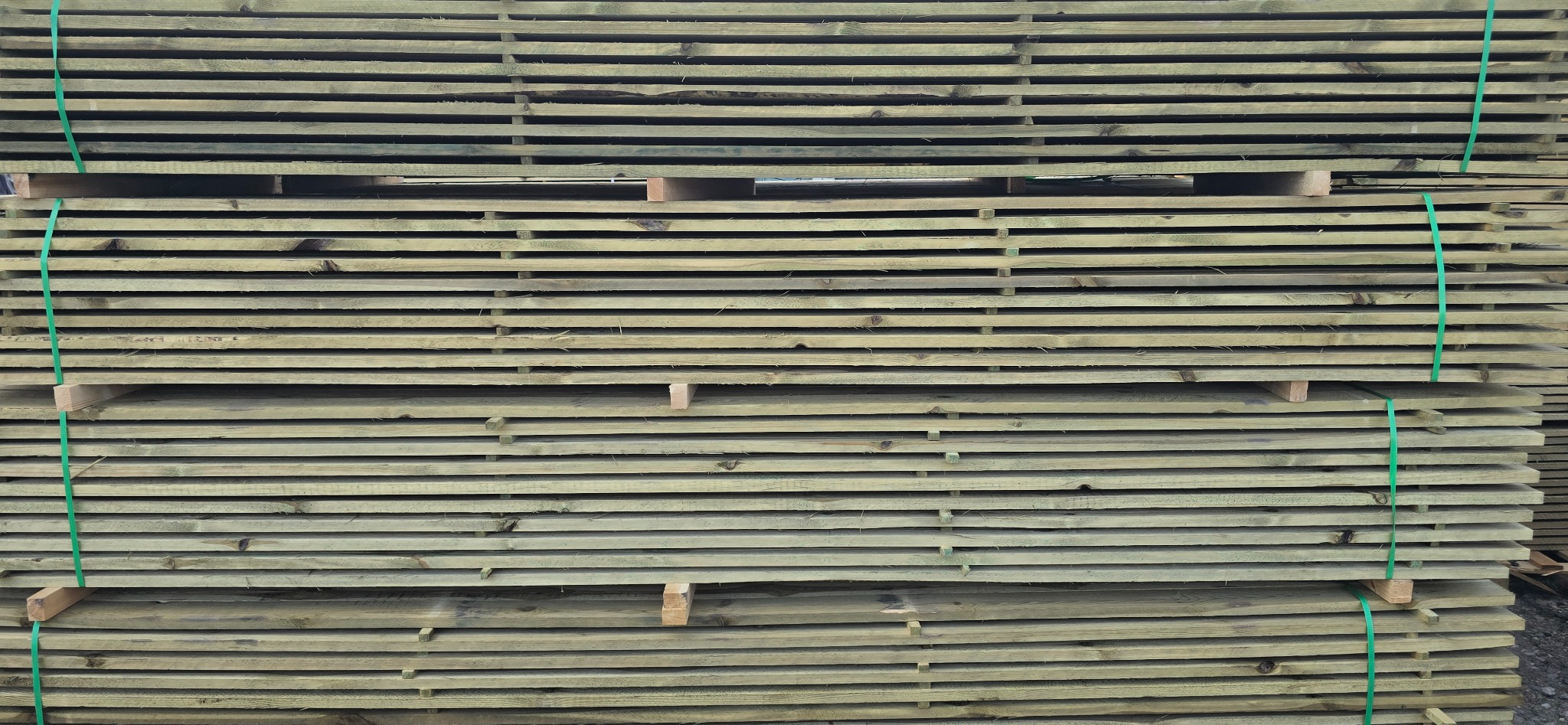 Stos desek o zielonkawym odcieniu, ułożonych równo jedna na drugiej i spiętych zielonymi taśmami. Widoczne przekładki między warstwami drewna.