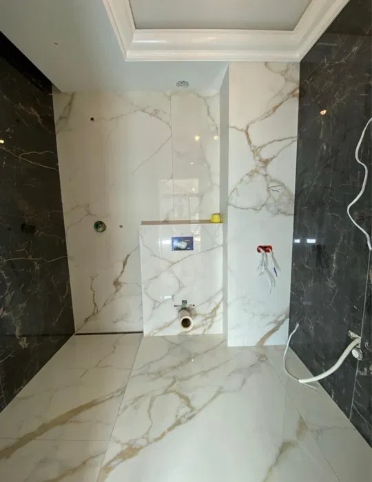 Wnętrze łazienki w trakcie wykańczania, z białymi i czarnymi płytkami imitującymi marmur z wyraźnymi złotymi żyłami na ścianach i podłodze. Widoczne elementy instalacji sanitarnej, takie jak rura...