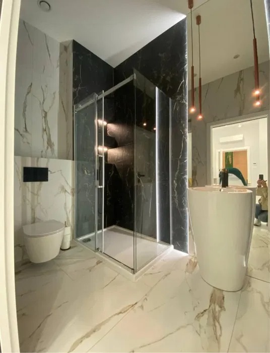 Nowoczesna łazienka z kabiną prysznicową narożną z czarnymi płytkami imitującymi marmur, białą umywalką wolnostojącą i toaletą podwieszaną, oświetlona wiszącymi lampami tubami w kolorze miedzianym.