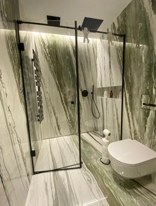 Nowoczesna kabina prysznicowa z czarną ramą i deszczownicą, biała toaleta wisząca, ściany i podłoga wyłożone marmurem w odcieniach bieli i zieleni, dekoracyjne figurki na kabinie.