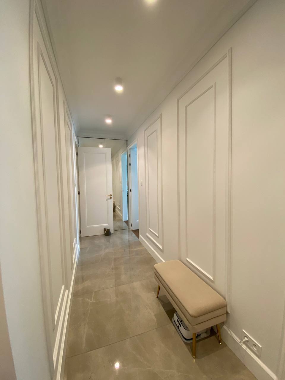 Długi korytarz z panelami ściennymi w kolorze złamanej bieli, polerowana podłoga z płytek imitujących marmur, beżowa ławka z złotymi nogami, oświetlenie punktowe sufitowe i lustro na końcu...