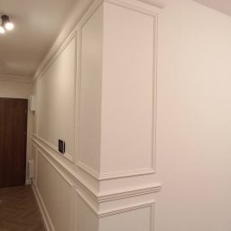 Stan Rem Bud - Białe ściany z ozdobnymi listwami i panelami, parkiet ułożony w jodełkę, widok z perspektywy narożnika, oświetlenie sufitowe.