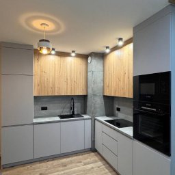 Stan Rem Bud - Nowoczesna kuchnia z szarymi szafkami dolnymi i drewnopodobnymi górnymi, czarnym zlewem i płytą indukcyjną, oświetlona wiszącą lampą i punktowymi reflektorami, z podłogą imitującą drewno.
