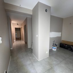 Stan Rem Bud - Korytarz w trakcie prac wykończeniowych: świeżo położone szare płytki podłogowe, ściany w odcieniach beżu, częściowo pomalowane, widoczna puszka z farbą i narzędzia w tle.