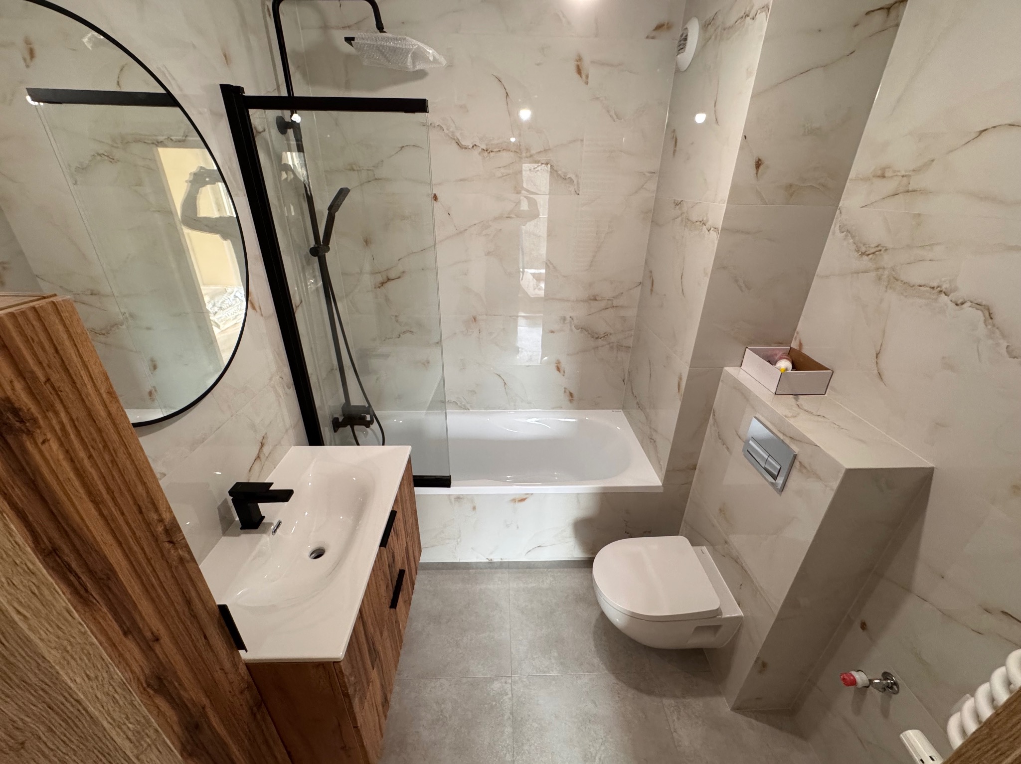 Nowoczesna łazienka z marmurowymi płytkami, drewnianą szafką pod umywalką z czarną armaturą, kabiną prysznicową typu walk-in z czarnymi ramami i owalnym lustrem.