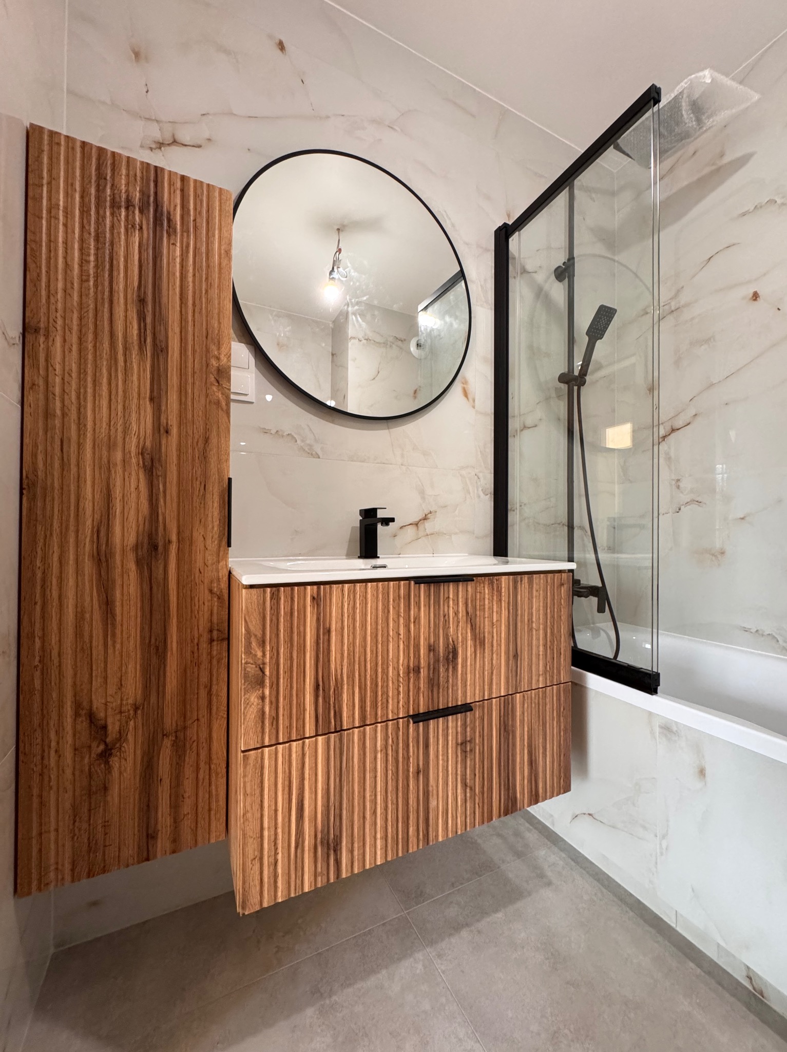 Nowoczesna łazienka z drewnianą szafką pod umywalką z frezowanymi frontami, okrągłym lustrem w czarnej ramie i kabiną prysznicową typu walk-in z czarnymi profilami.