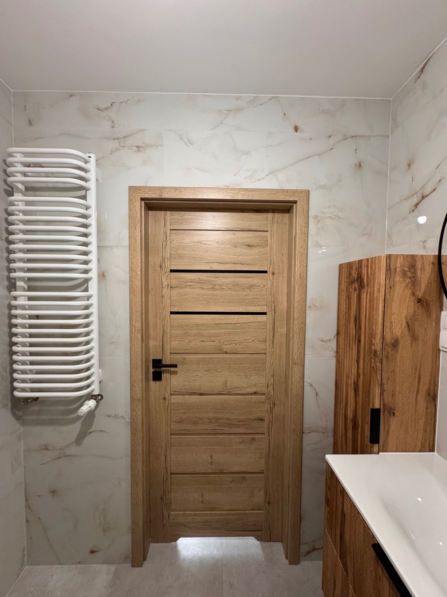 Nowoczesna łazienka z marmurowymi płytkami, drewnianą zabudową umywalki i drzwiami o naturalnym odcieniu drewna z poziomymi wstawkami, biały grzejnik drabinkowy.