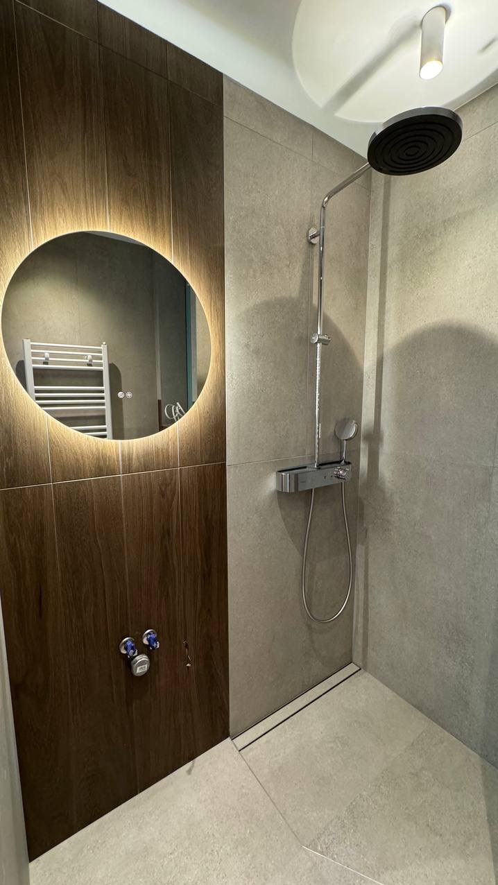 Nowoczesna kabina prysznicowa z deszczownicą, szarymi płytkami i podświetlanym lustrem owalnym, w którym odbija się drabinka na ręczniki.