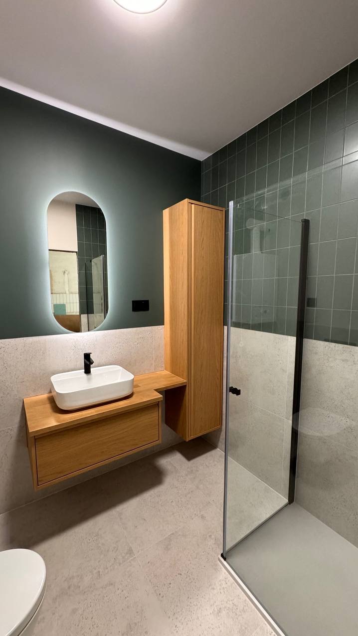 Nowoczesna łazienka z drewnianymi szafkami, podświetlanym lustrem owalnym, zielonymi płytkami i kabiną prysznicową ze szklaną ścianą.