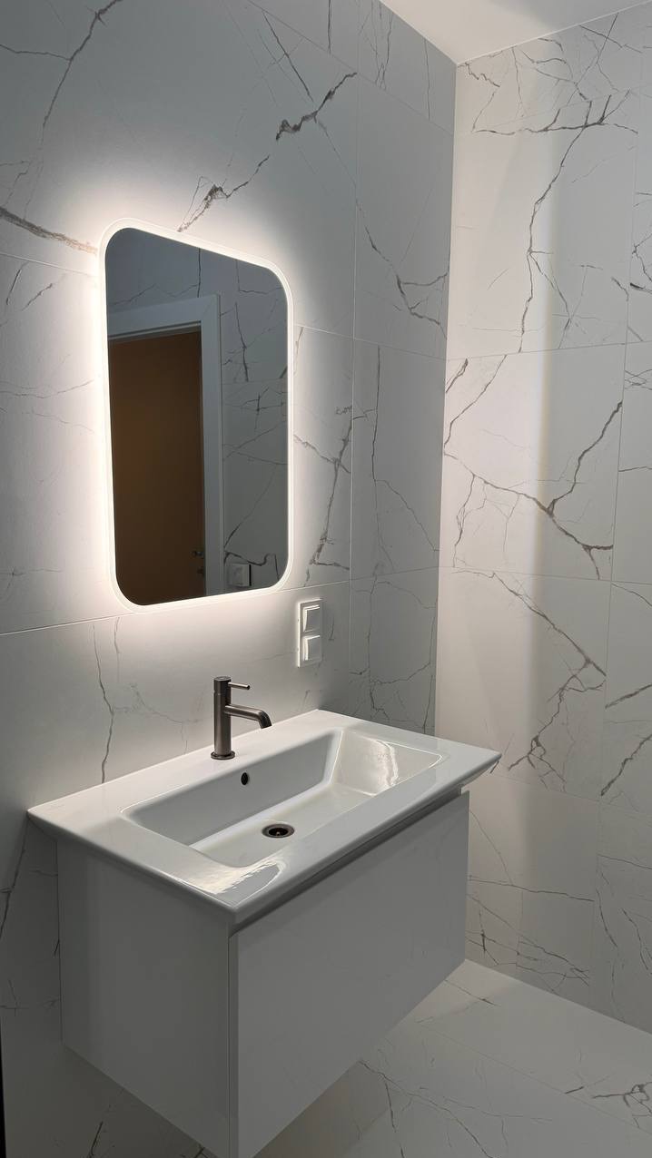 Nowoczesna łazienka z umywalką nablatową na szafce, podświetlanym lustrem i płytkami imitującymi marmur na ścianach i podłodze.
