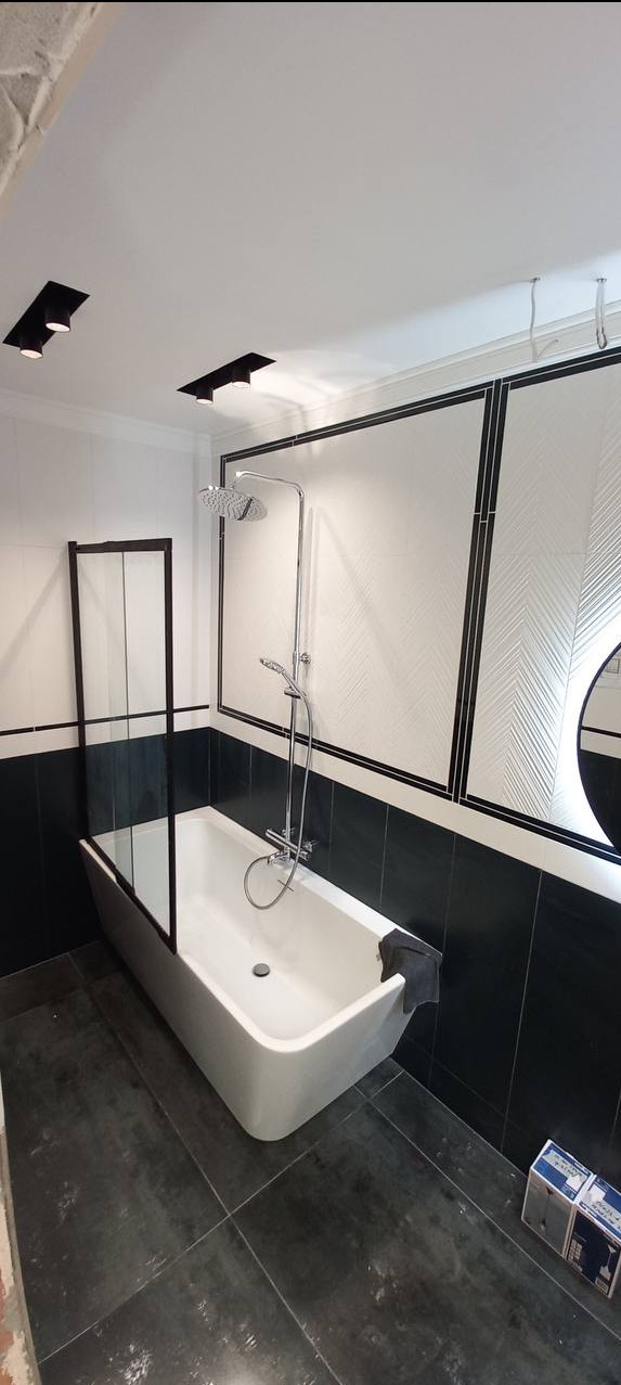 Nowoczesna łazienka w czerni i bieli: wanna z parawanem, deszczownica, płytki chevron na ścianie, ciemna podłoga, oświetlenie punktowe.