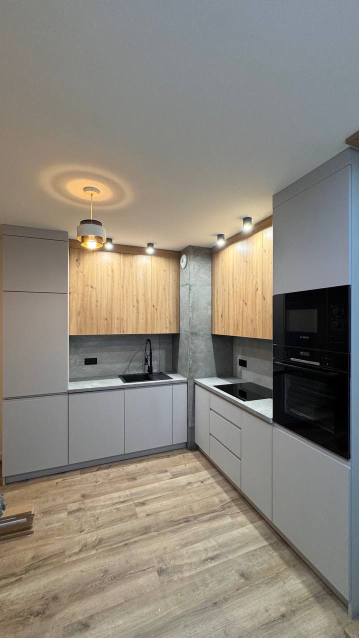 Nowoczesna kuchnia z szarymi szafkami dolnymi i drewnopodobnymi górnymi, czarnym zlewem i płytą indukcyjną, oświetlona wiszącą lampą i punktowymi reflektorami, z podłogą imitującą drewno.