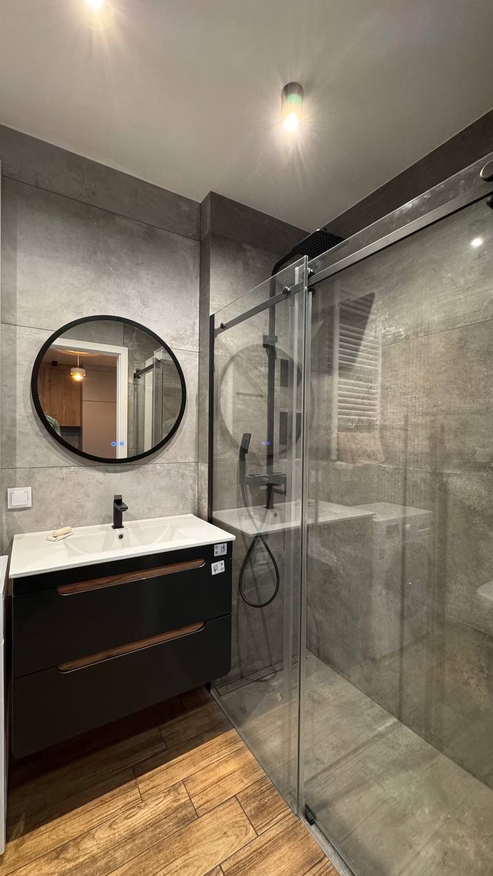 Nowoczesna łazienka z kabiną prysznicową typu walk-in, okrągłym lustrem z podświetleniem i umywalką nablatową z czarną armaturą, wykończona płytkami imitującymi beton i drewnopodobną podłogą.