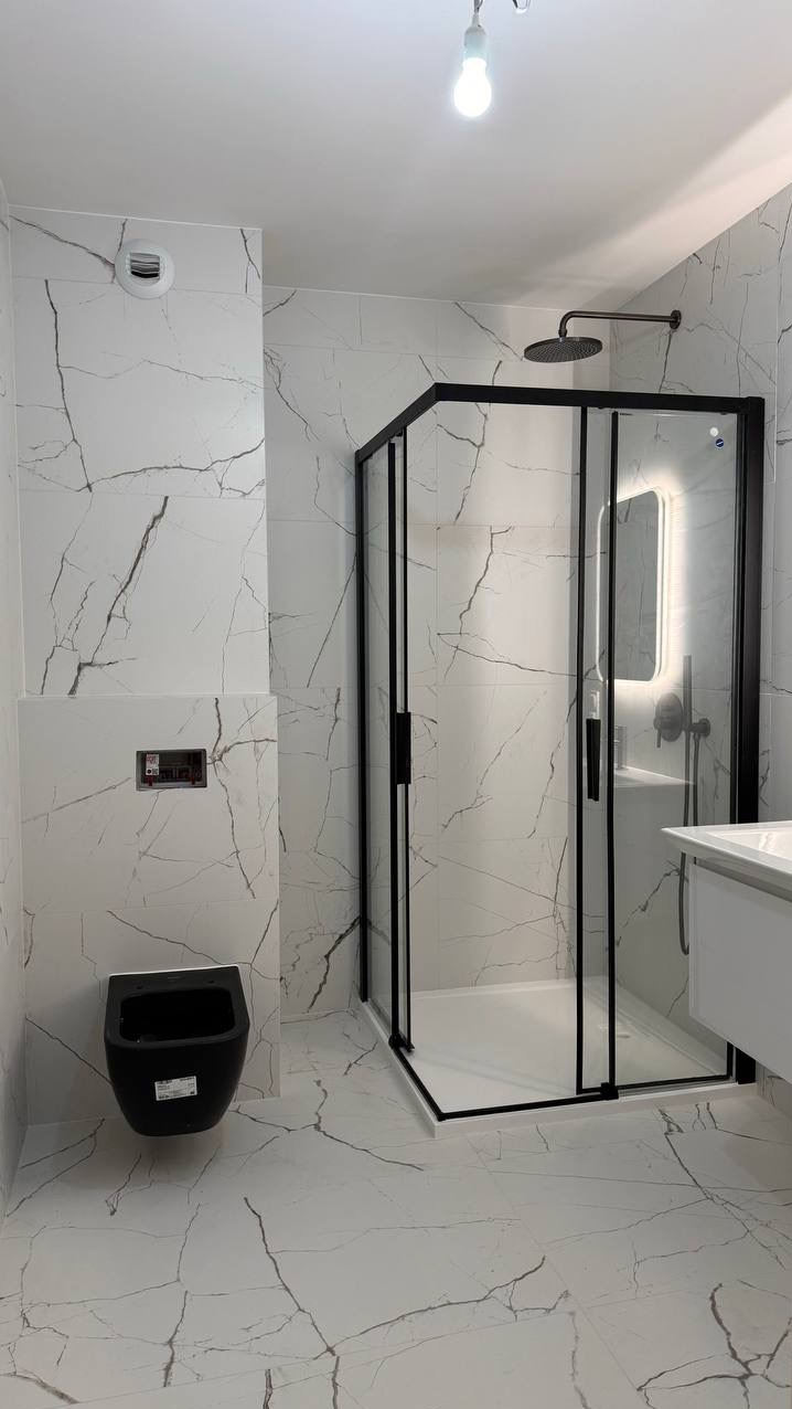 Nowoczesna łazienka w Krakowie wykończona białymi płytkami imitującymi marmur, z czarną kabiną prysznicową typu walk-in, podwieszaną miską WC i oświetlonym lustrem.