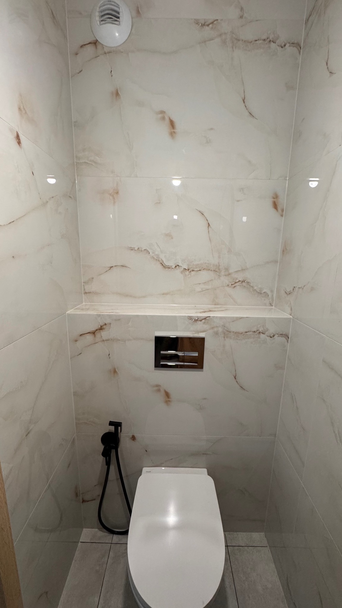 Wykończona łazienka z toaletą wiszącą, bidetką w kolorze czarnym i płytkami imitującymi marmur z brązowymi żyłami na ścianach i półką.