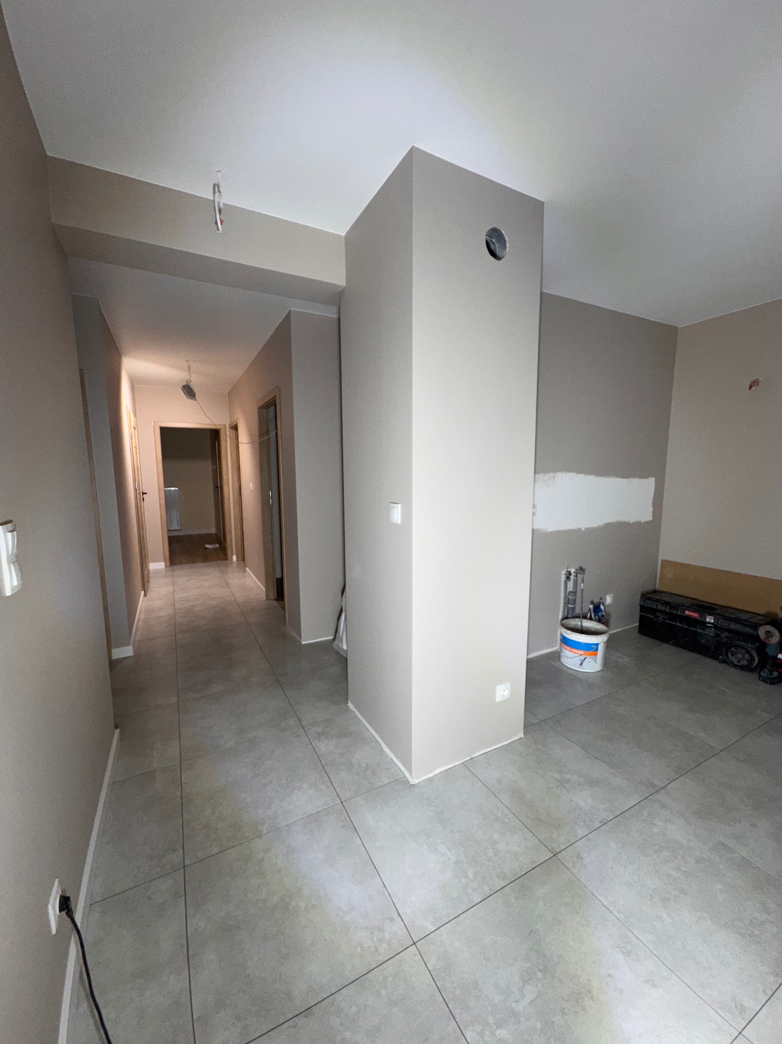 Korytarz w trakcie prac wykończeniowych: świeżo położone szare płytki podłogowe, ściany w odcieniach beżu, częściowo pomalowane, widoczna puszka z farbą i narzędzia w tle.