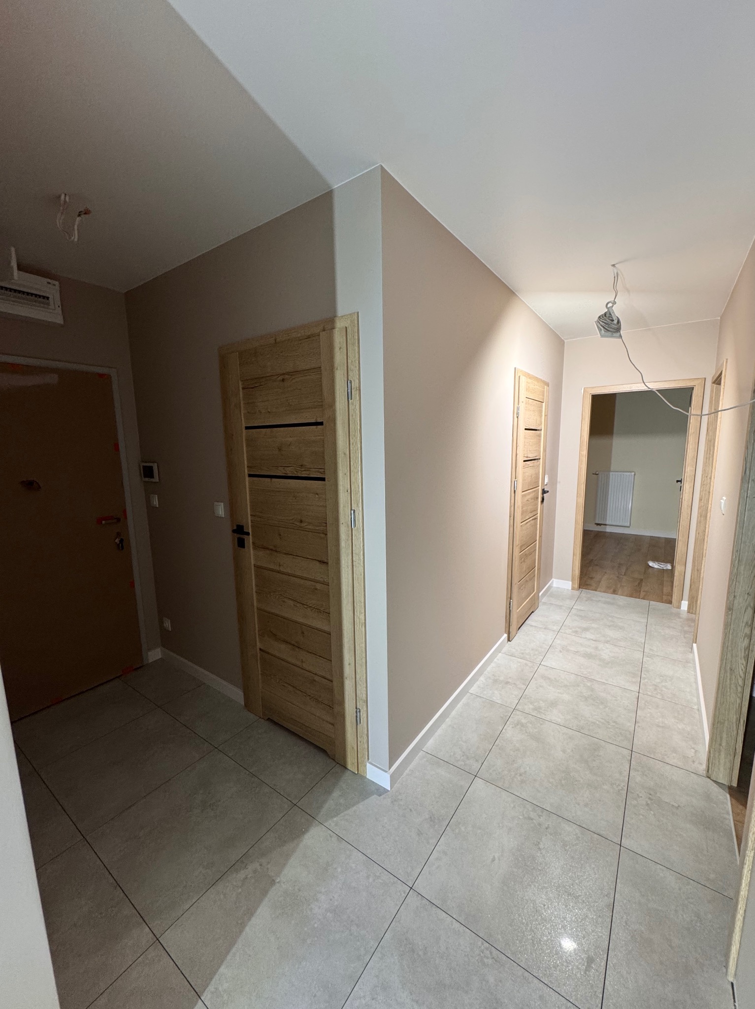Korytarz po remoncie z beżowymi ścianami, drewnianymi drzwiami i szarą podłogą z płytek; widoczne niedokończone podłączenie oświetlenia na suficie.