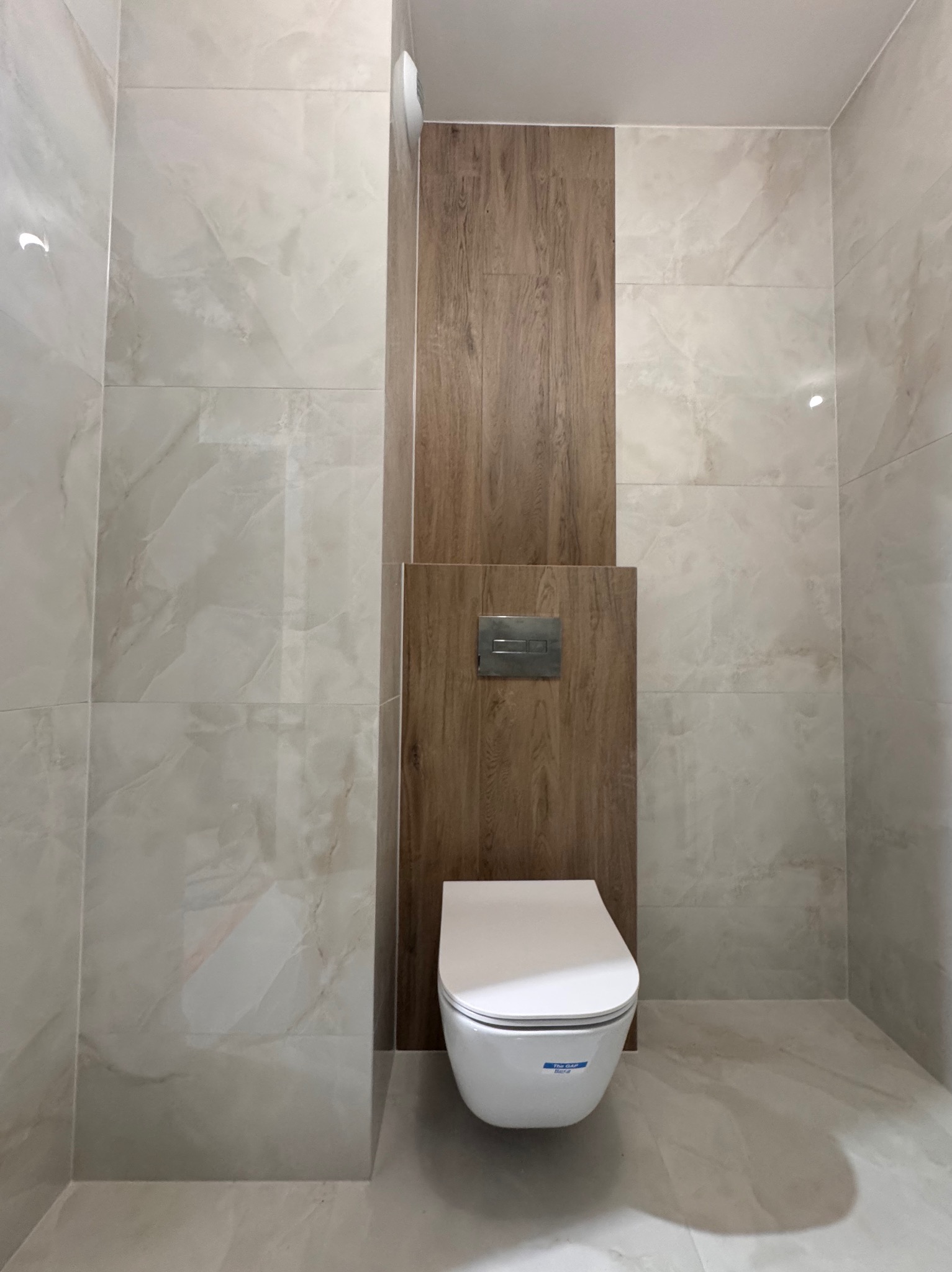 Wykończona łazienka z podwieszaną toaletą, płytkami imitującymi marmur i drewnopodobną zabudową stelaża, widoczne chromowane przyciski spłukiwania.