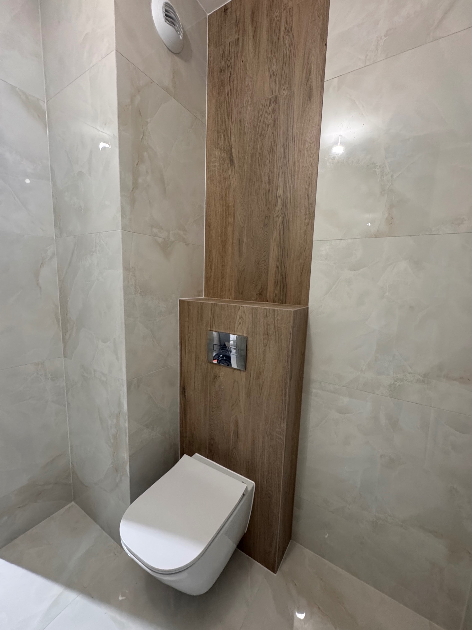 Nowoczesna toaleta podwieszana z drewnianą obudową stelaża, marmurkowe płytki na ścianach i podłodze, srebrny przycisk spłukiwania.