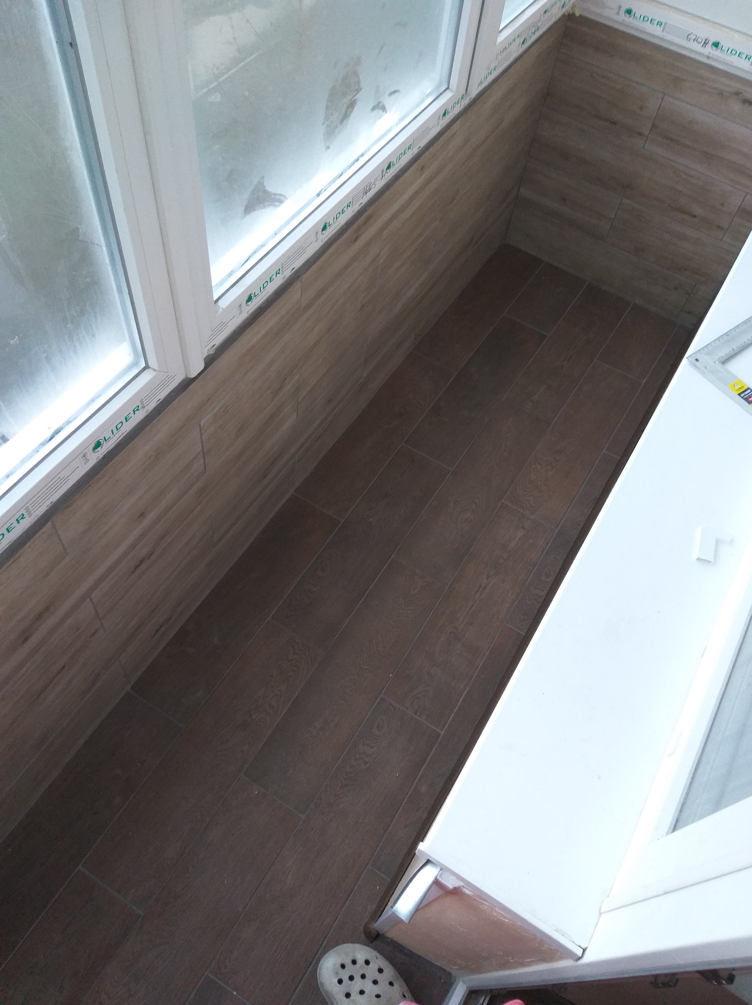 Wykończony balkon z brązowymi płytkami imitującymi drewno na podłodze i ścianie, biała rama okienna z widocznym profilem 'Lider' i fragment buta w rogu kadru.