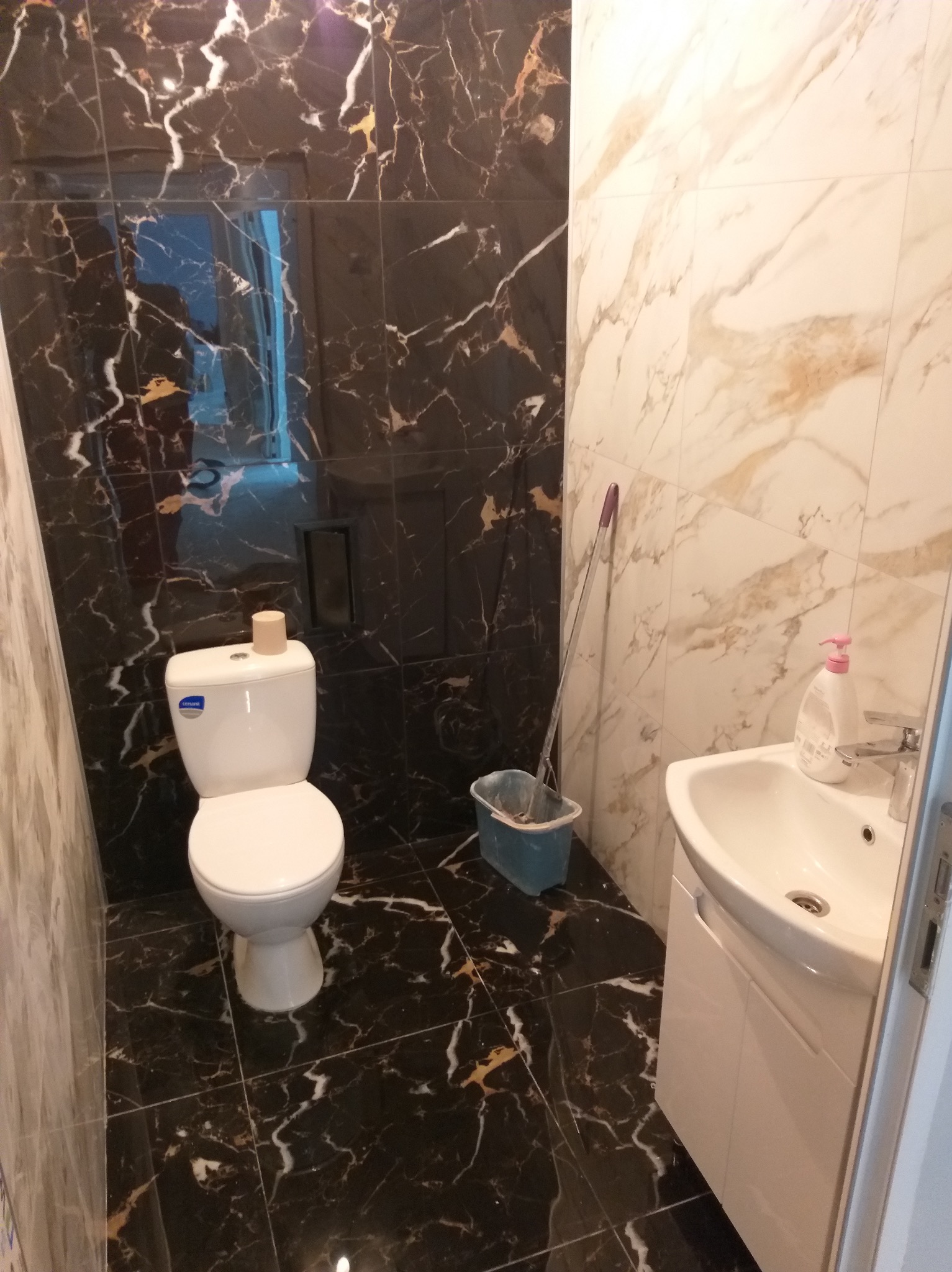 Wykończona łazienka z czarno-złotymi marmurowymi płytkami na ścianach i podłodze, białą armaturą i mopem opartym o ścianę.