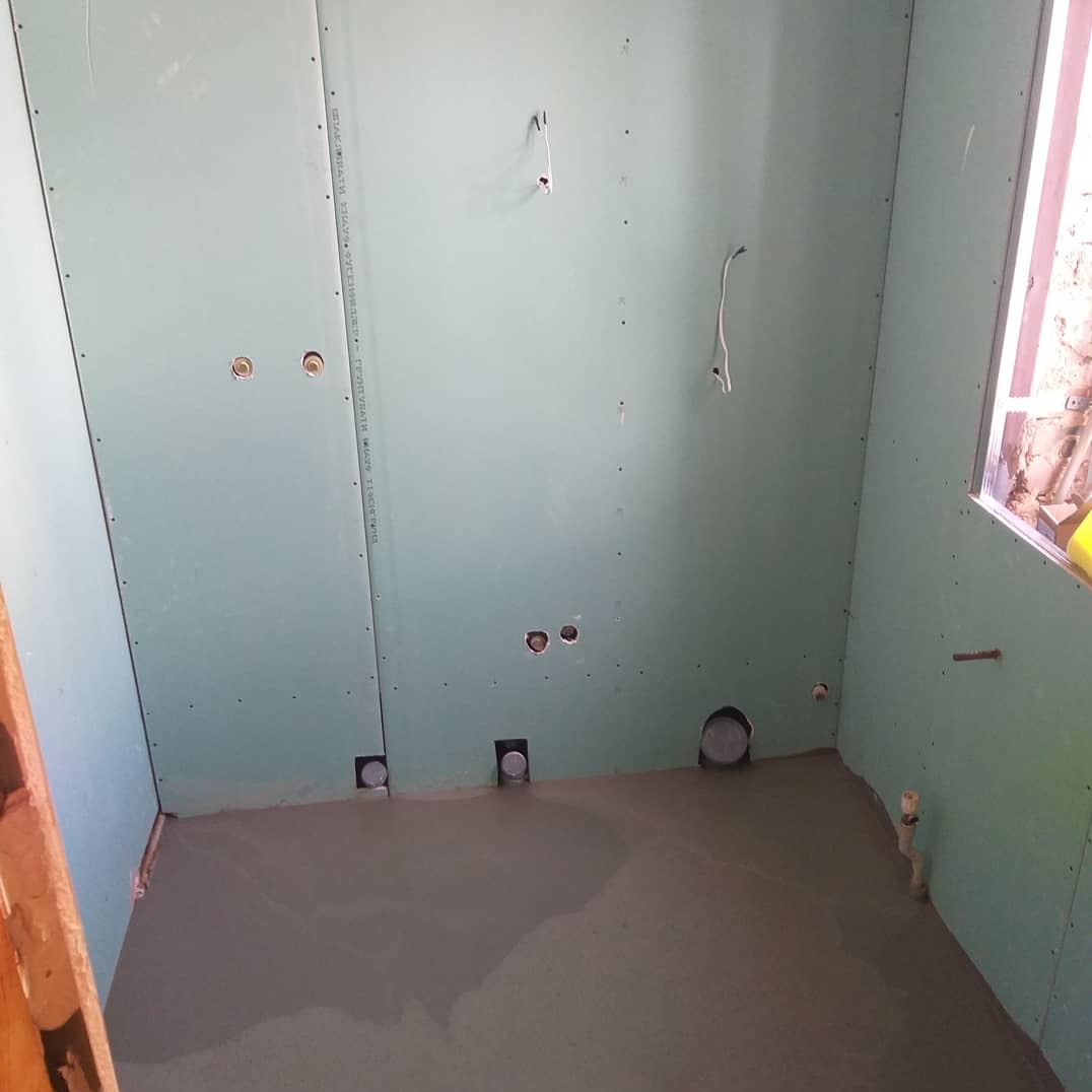 Łazienka w trakcie remontu: świeżo wylana wylewka podłogowa, ściany obłożone zielonymi płytami gipsowo-kartonowymi, widoczne otwory na instalacje.