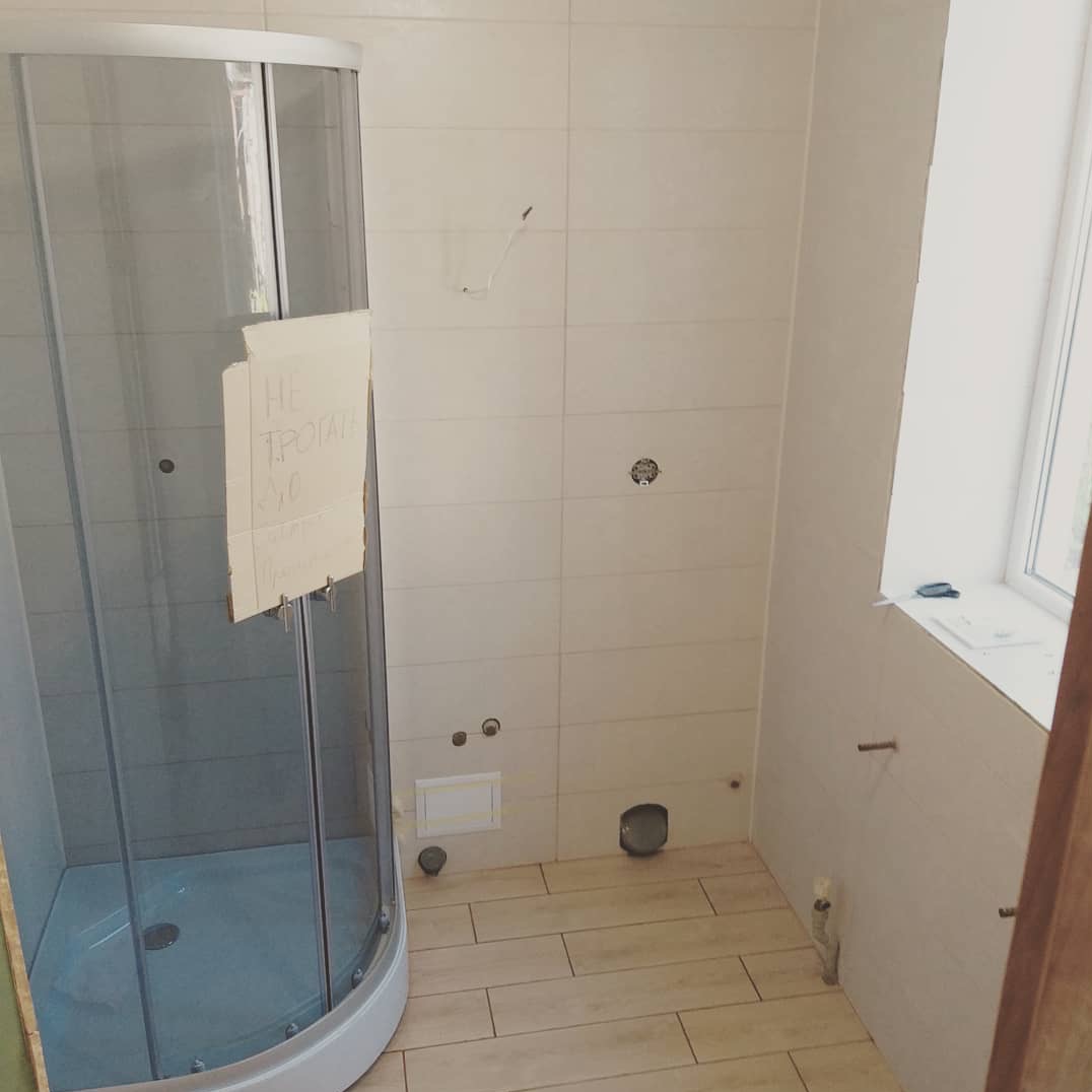 Wnętrze łazienki w trakcie remontu: kabina prysznicowa z kartką 'Nie dotykać' zawieszoną na drzwiach, jasne płytki na ścianach, drewnopodobna podłoga, widoczne otwory na instalacje.