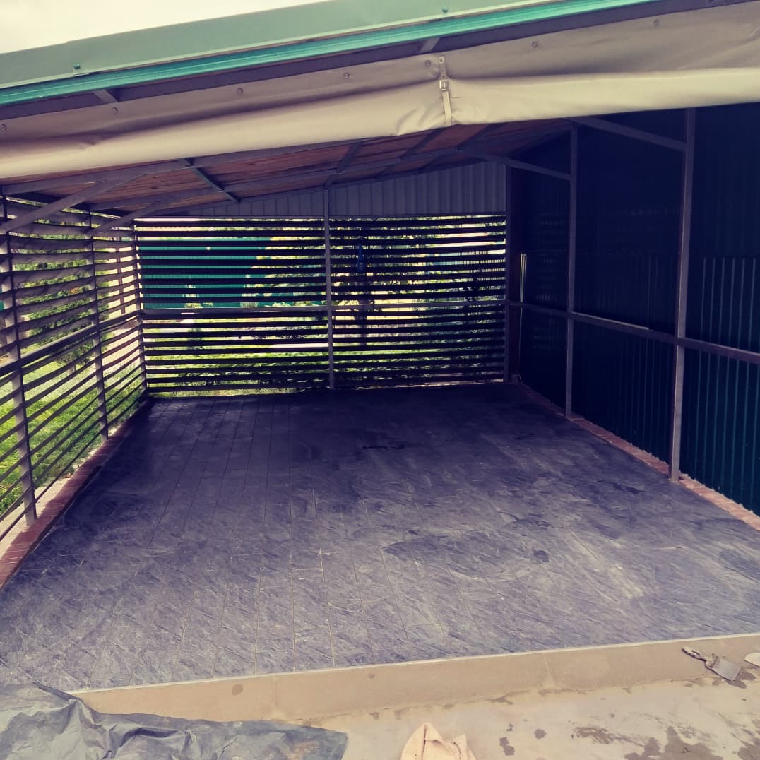 Zadaszone patio z drewnianą konstrukcją i szarą podłogą przygotowaną pod wykończenie, widok z zewnątrz przez drewniane żaluzje.