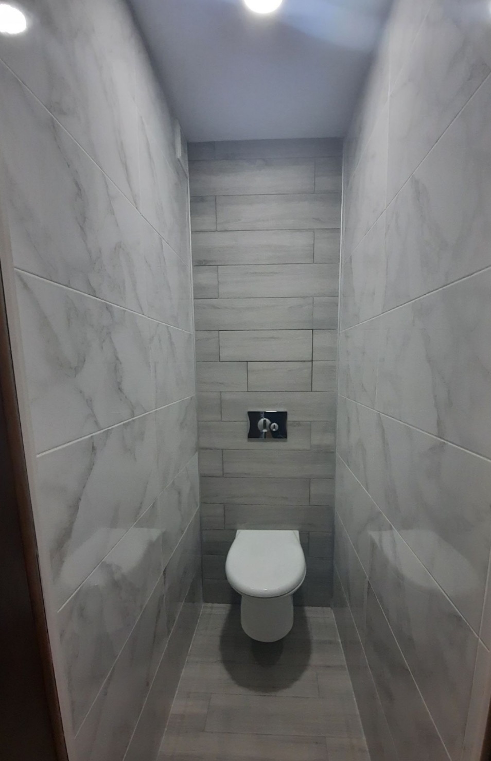 Wąska toaleta wykończona płytkami imitującymi marmur na ścianach bocznych i szarymi płytkami drewnopodobnymi na ścianie frontowej, z podtynkowym panelem spłukującym i wiszącą miską WC.