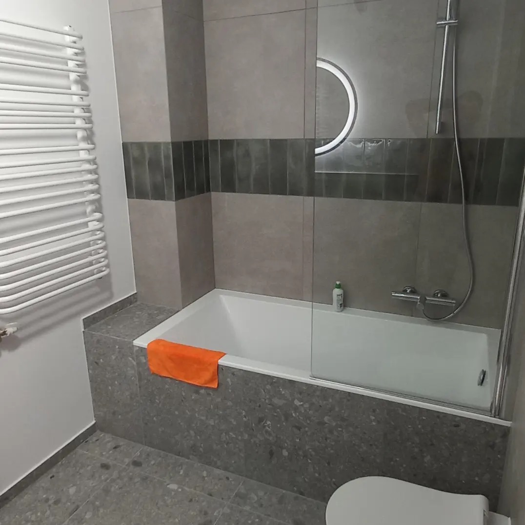 Nowoczesna łazienka z wanną obudowaną szarą mozaiką, szklanym parawanem nawannowym, okrągłym podświetlanym lustrem i grzejnikiem drabinkowym; pomarańczowy ręcznik na krawędzi wanny.