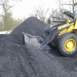 Żółta ładowarka XCMG Europa z czarną łyżką nabiera górę czarnego, błyszczącego węgla na placu składowym, z drzewami w tle.