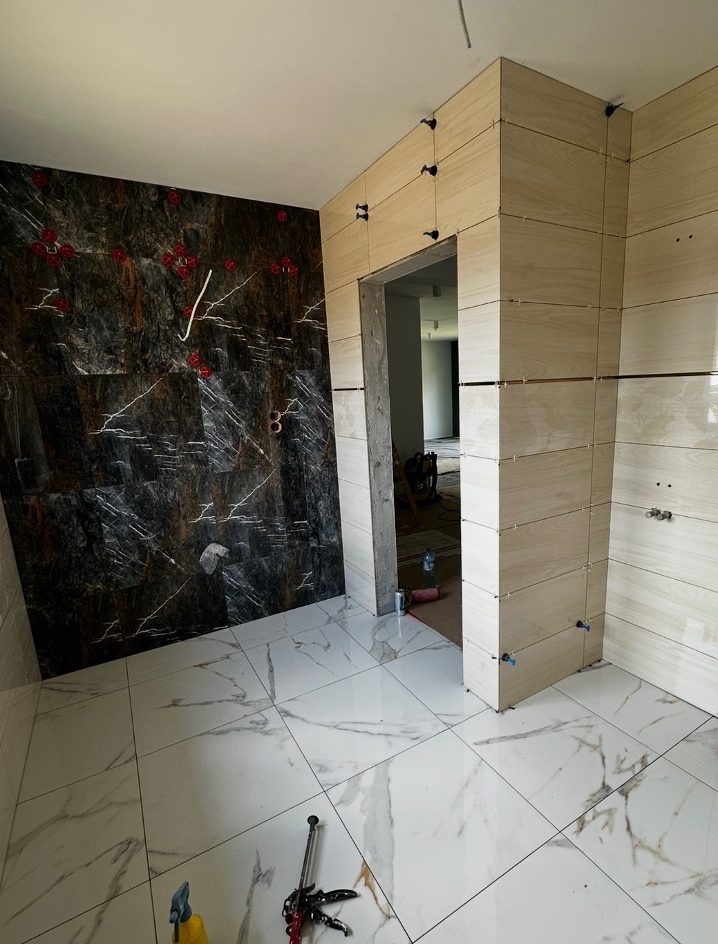 Układanie płytek w łazience: marmurowe płytki podłogowe i ścienne w odcieniach beżu oraz czerni. Widoczne narzędzia glazurnicze i system poziomowania płytek.