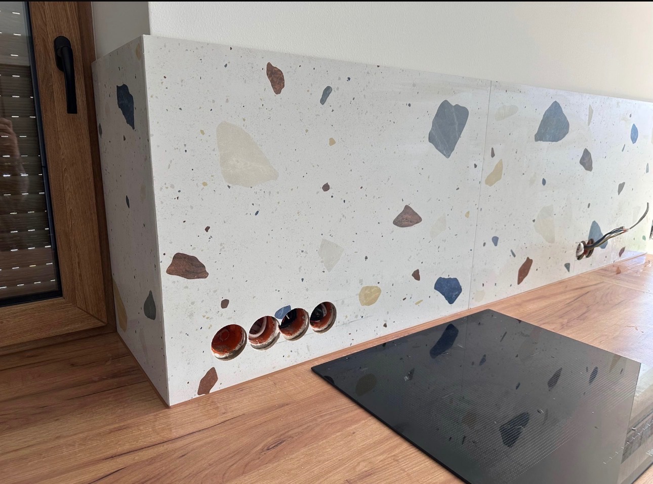 Ściana z płytek lastryko w kuchni, z wywierconymi otworami na gniazdka, obok płyta indukcyjna. Widoczny fragment okna z roletami. Ujęcie z niskiego kąta.