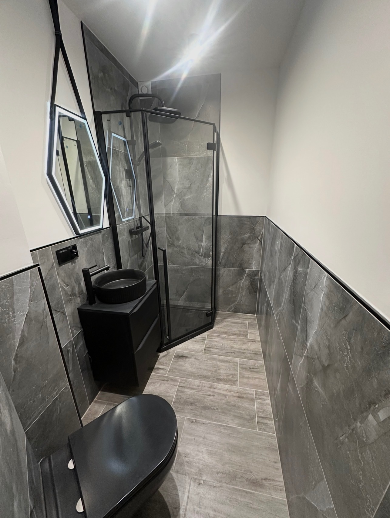 Nowoczesna łazienka w szarościach z czarnymi akcentami: kabina prysznicowa, umywalka na szafce i toaleta. Uwagę przykuwa heksagonalne lustro z podświetleniem LED.