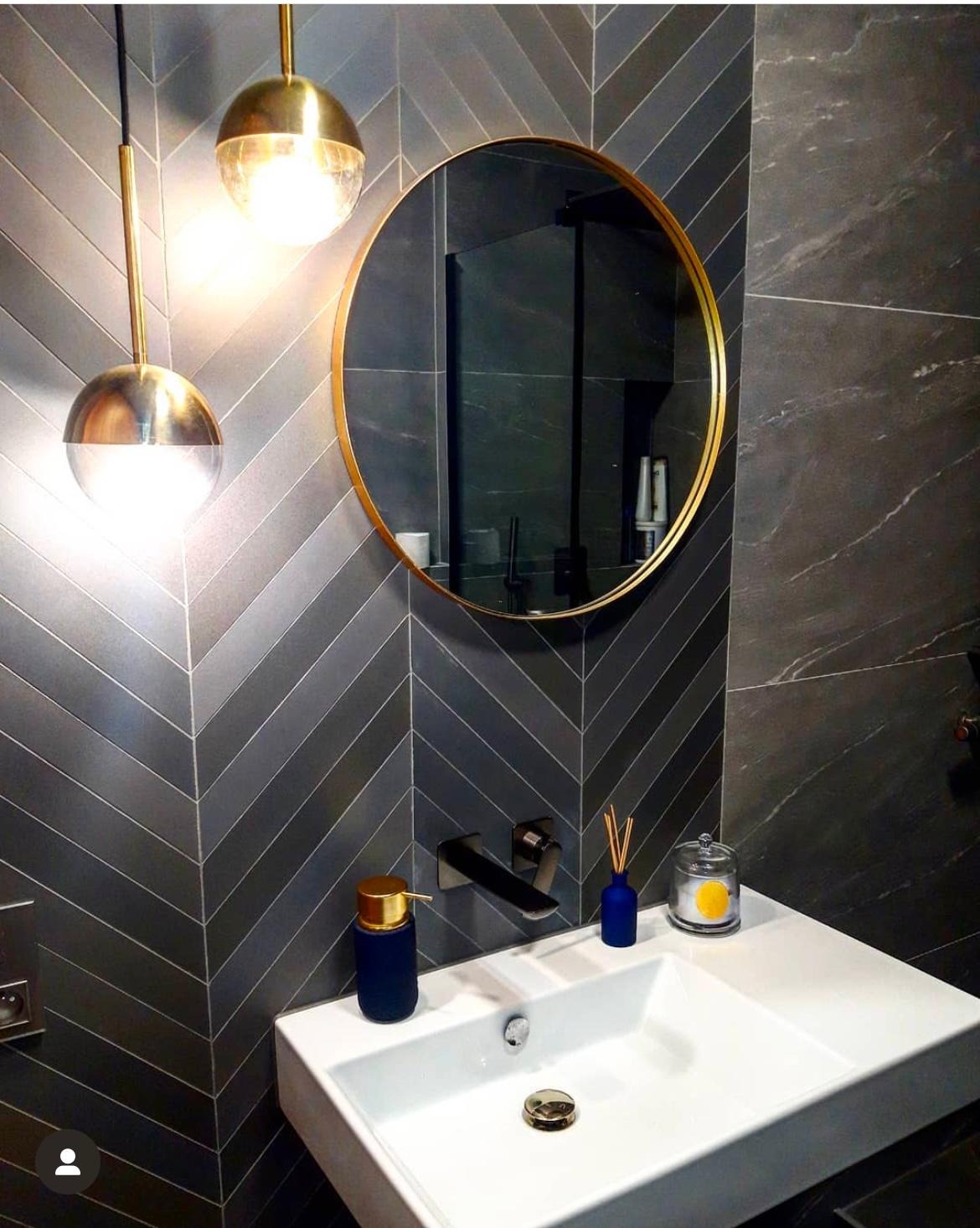 Nowoczesna łazienka z szarymi płytkami ułożonymi w jodełkę, okrągłe lustro w złotej ramie, biała umywalka z czarną baterią, dekoracyjne lampy wiszące w kształcie kul.