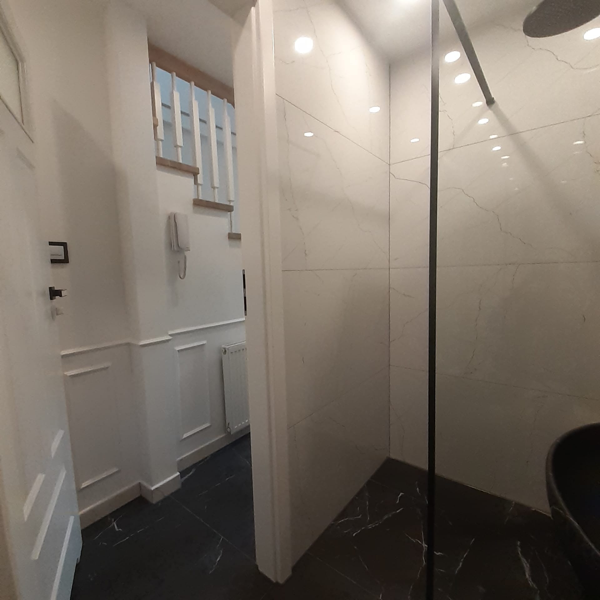 Wykończona łazienka z czarną armaturą i marmurowymi płytkami, widok na przedpokój z białymi panelami ściennymi i schodami.