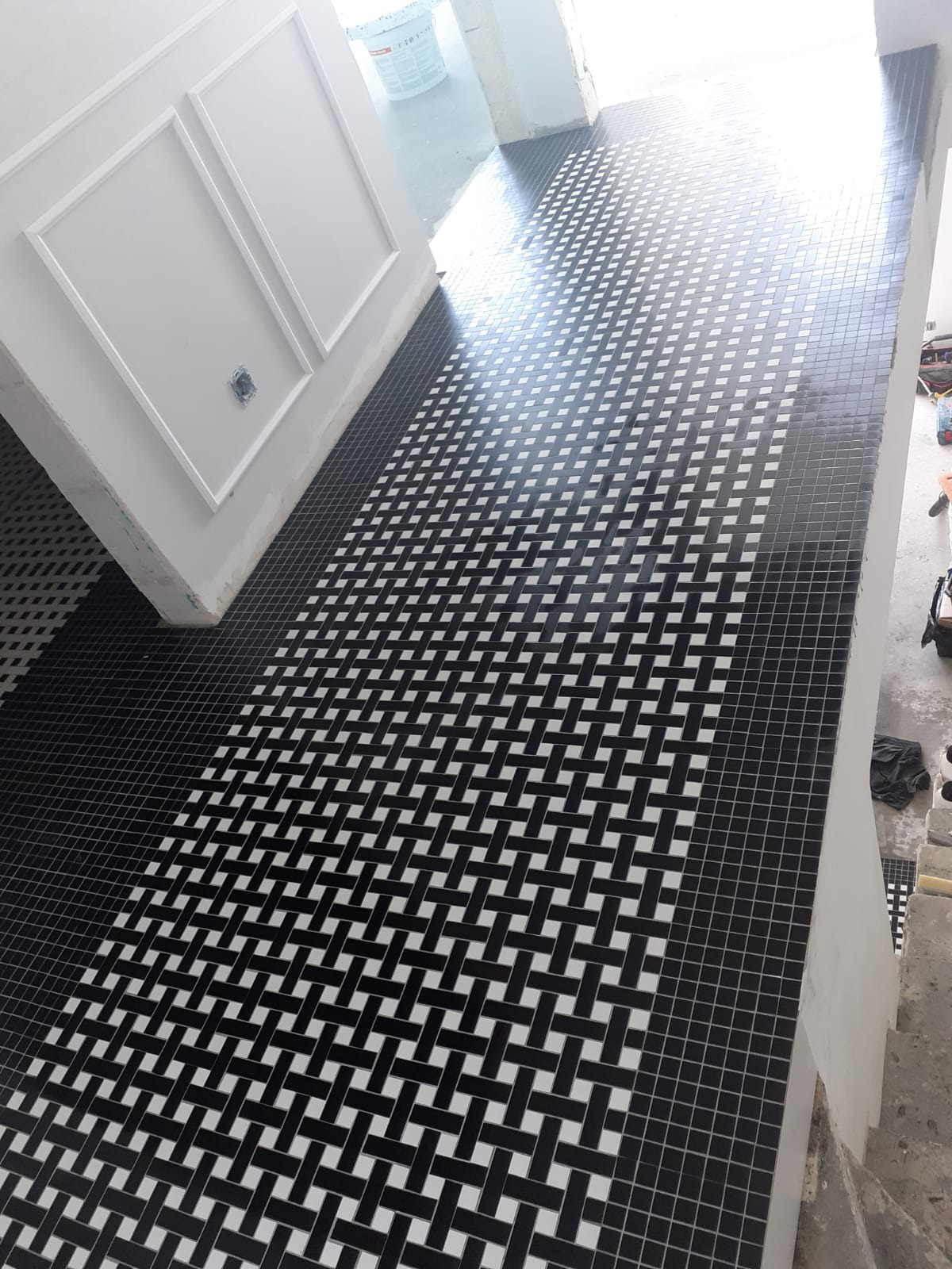 Ukończona podłoga z czarno-białej mozaiki ułożonej we wzór koszykowy, z widocznymi białymi listwami dekoracyjnymi na ścianie i wiaderkiem z zaprawą w tle.