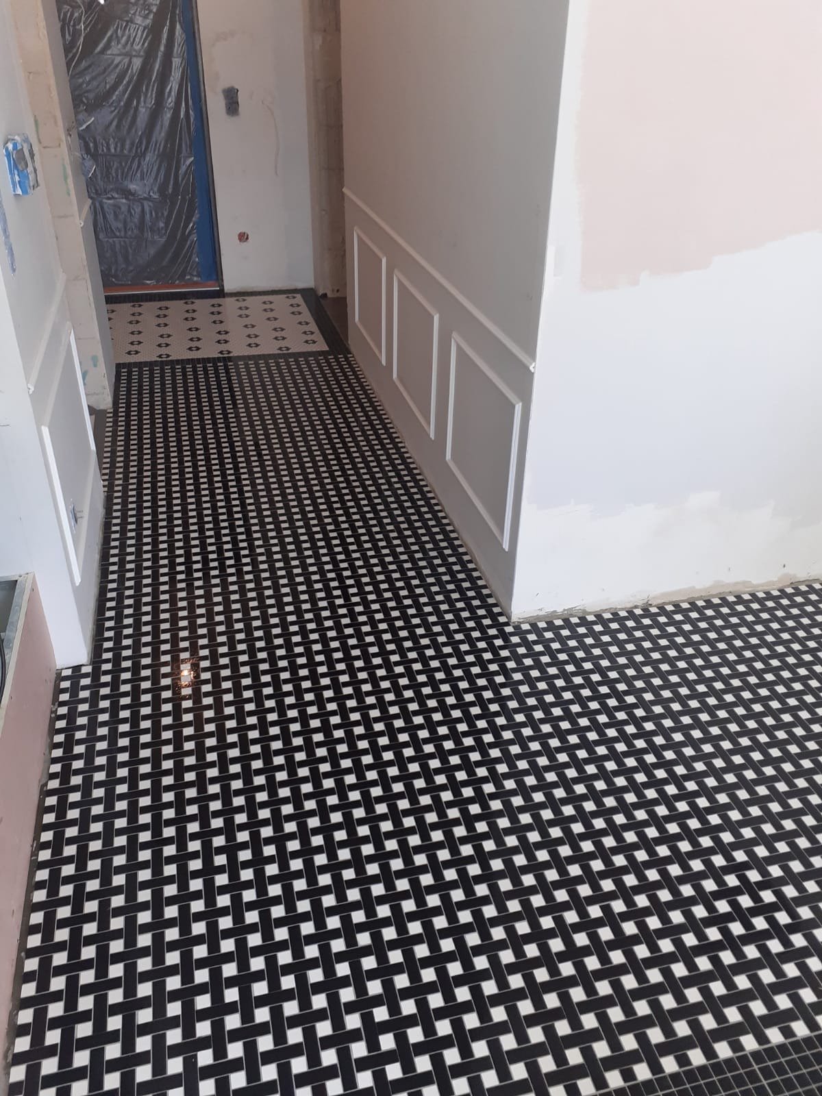 Ułożona czarno-biała mozaika podłogowa w wąskim korytarzu, widoczne listwy przypodłogowe i fragment ściany z dekoracyjnymi panelami ściennymi, zabezpieczone drzwi w tle.