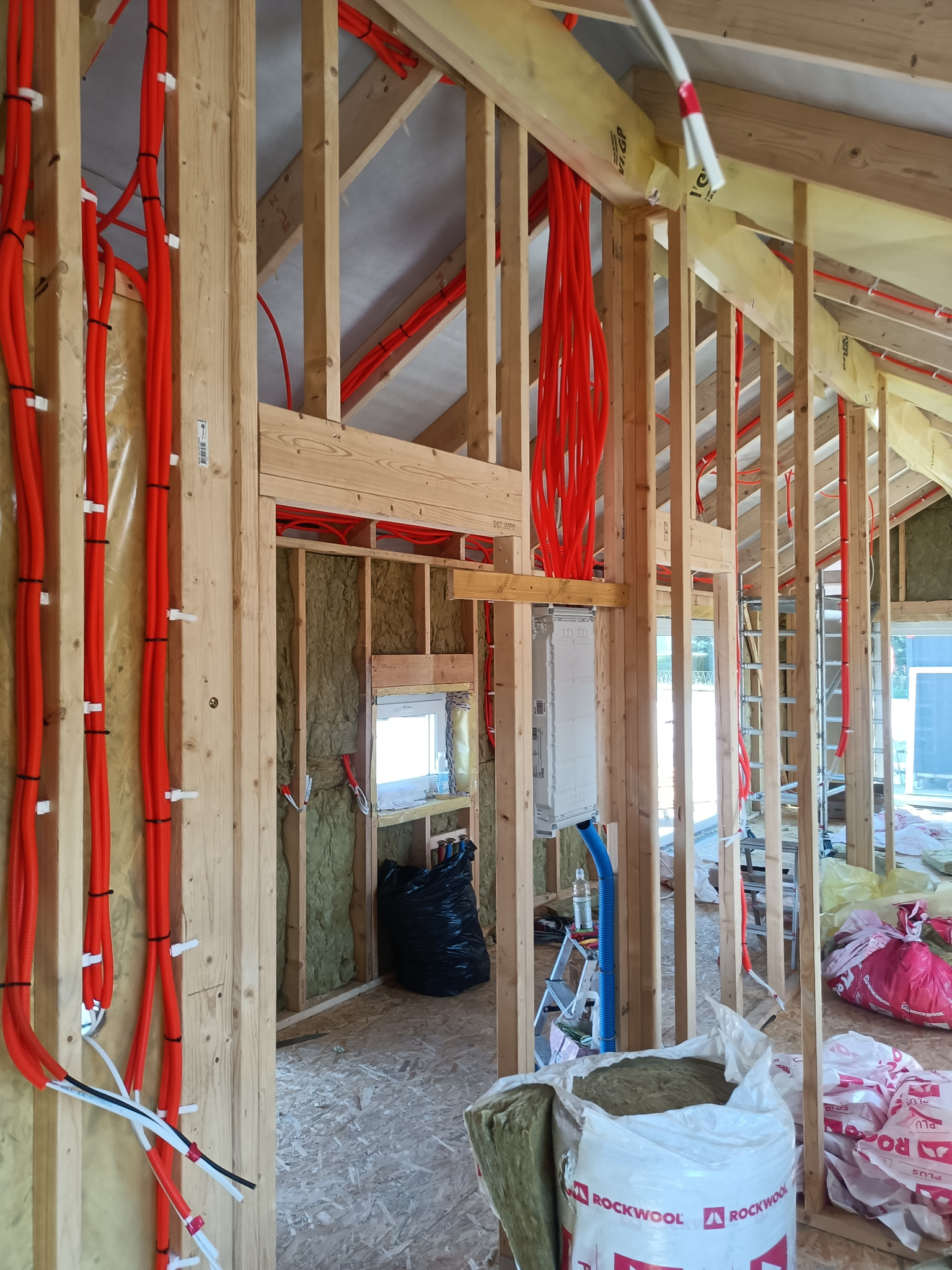 Wnętrze domu w trakcie budowy, widoczne drewniane szkielety ścian z poprowadzonymi czerwonymi rurami instalacji elektrycznej, częściowo ocieplone wełną mineralną, z rozdzielnicą elektryczną...