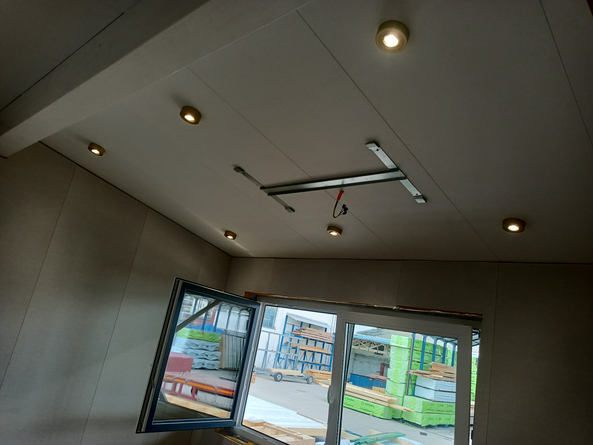 Wnętrze pomieszczenia z zamontowanymi okrągłymi lampami sufitowymi, widoczna szyna montażowa i otwarte okno z widokiem na plac budowy z materiałami budowlanymi.