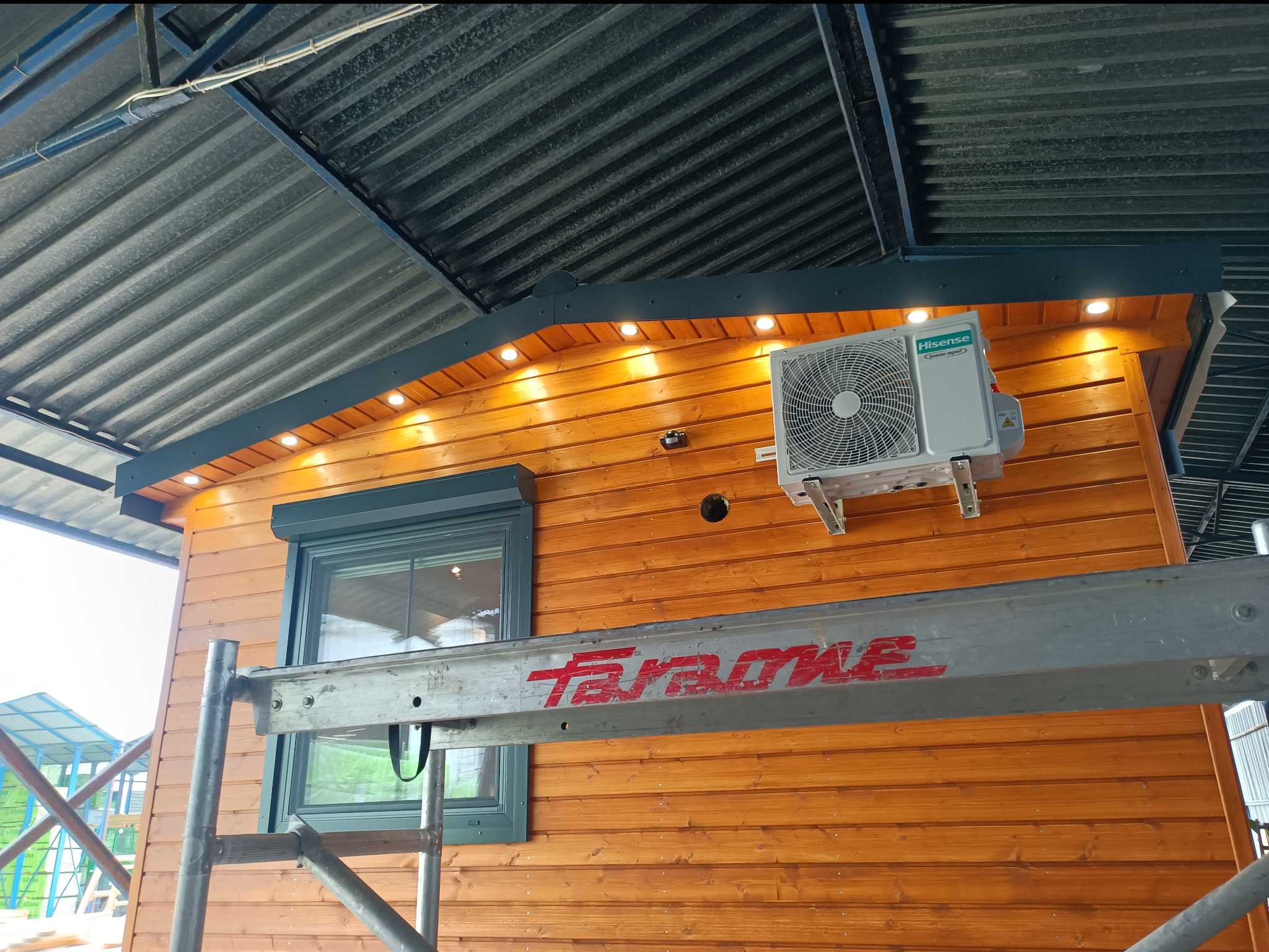 Klimatyzator Hisense zamontowany na drewnianej elewacji budynku z widoczną instalacją elektryczną i oświetleniem LED, widok z rusztowania.
