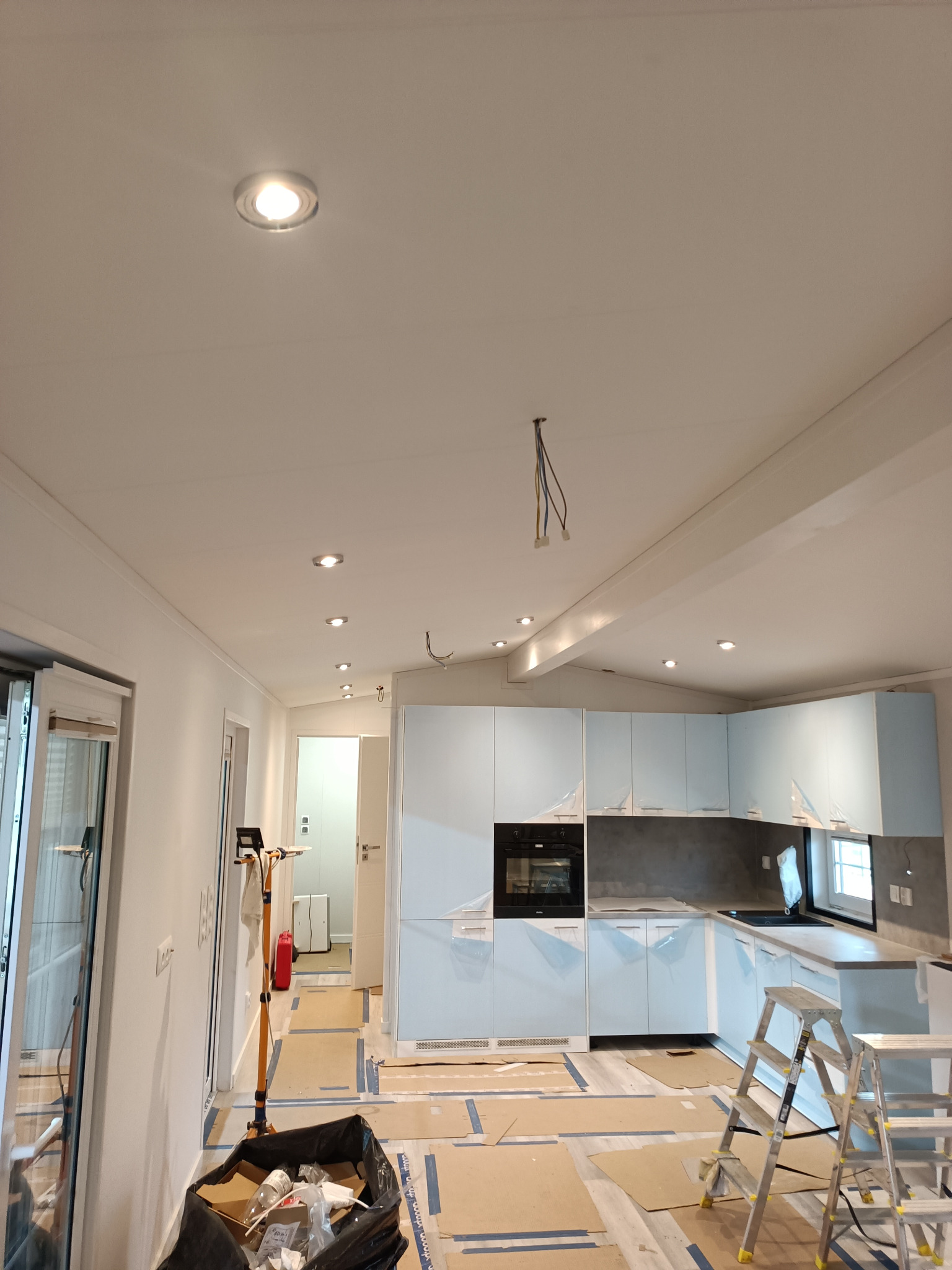 Wnętrze w trakcie remontu: zamontowane oświetlenie punktowe w suficie, częściowo zamontowana kuchnia z jasnoniebieskimi frontami, kartony zabezpieczające podłogę, widoczne kable wystające z sufitu.